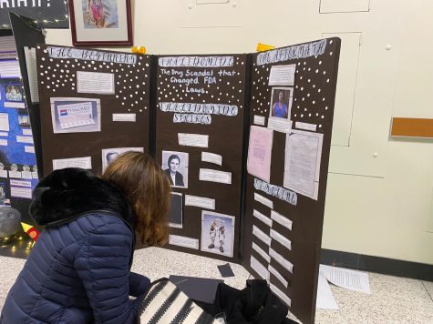 A junior puts finishing touches on her project about a federal drug scandal. She had mini pill bottles and white dots representing the drugs.